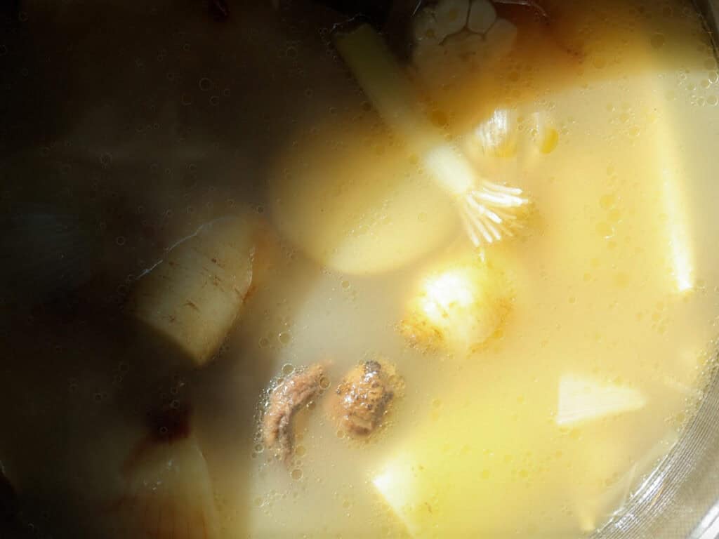 Close-up of a simmering soup with visible ingredients, including potatoes, onions, scallions, and other vegetables, in a rich, light-colored broth, partially lit by sunlight.